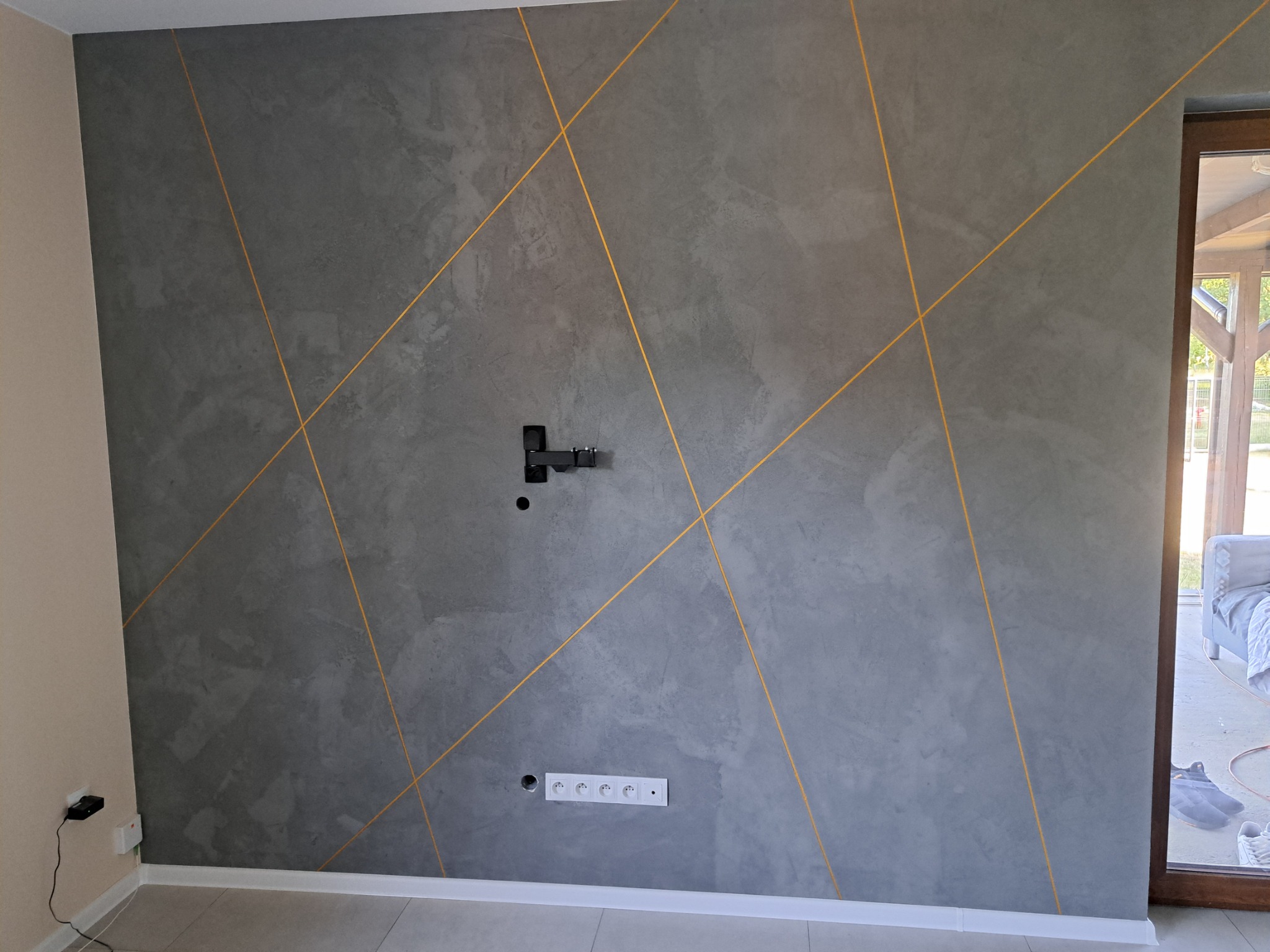 Ściana w salonie z szarą dekoracyjną fakturą i złotymi liniami, widoczne gniazdka elektryczne i uchwyt na telewizor.