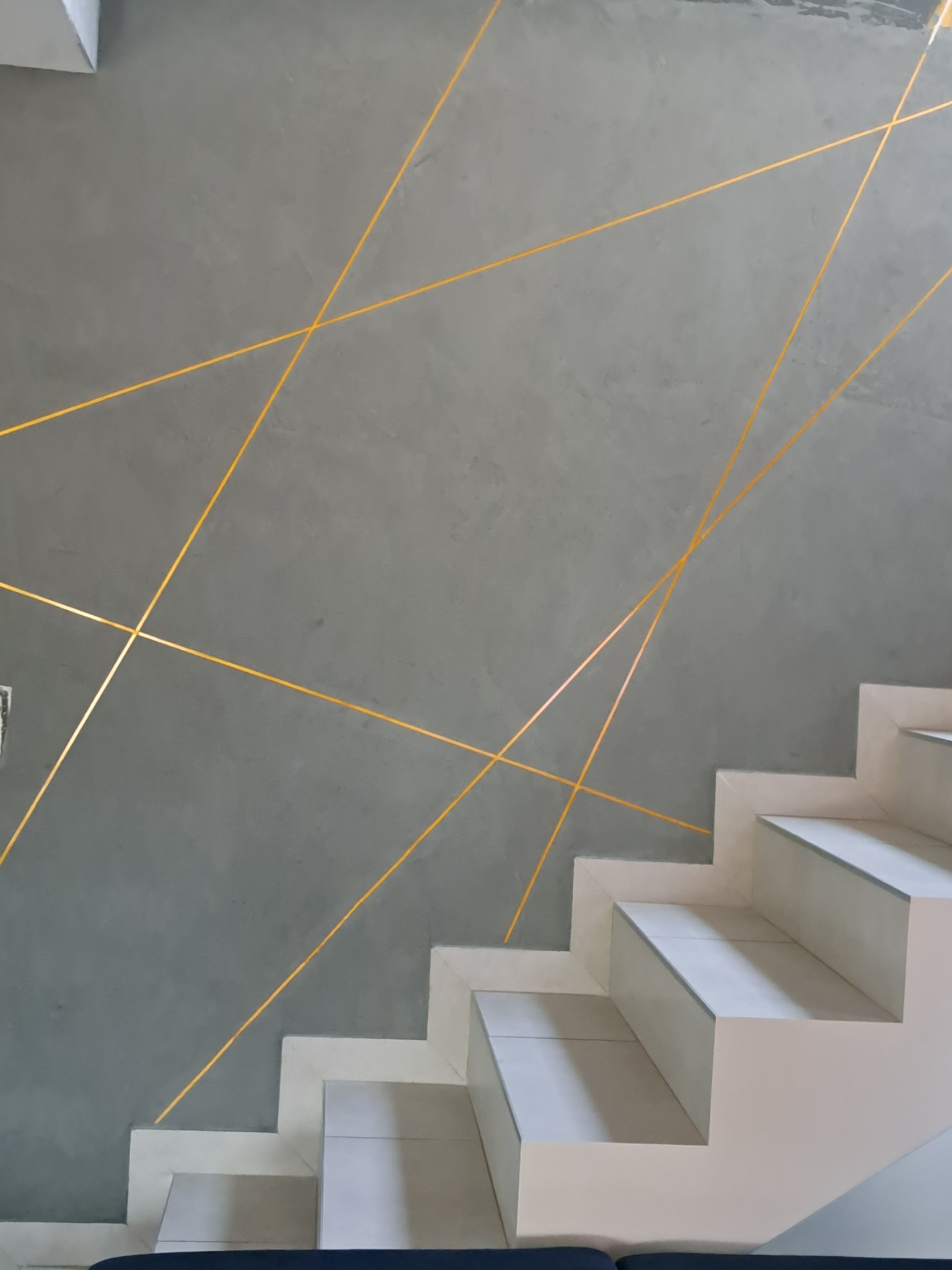 Szara ściana ze złotymi liniami tworzącymi geometryczny wzór, przylegająca do białych schodów z szarymi akcentami.