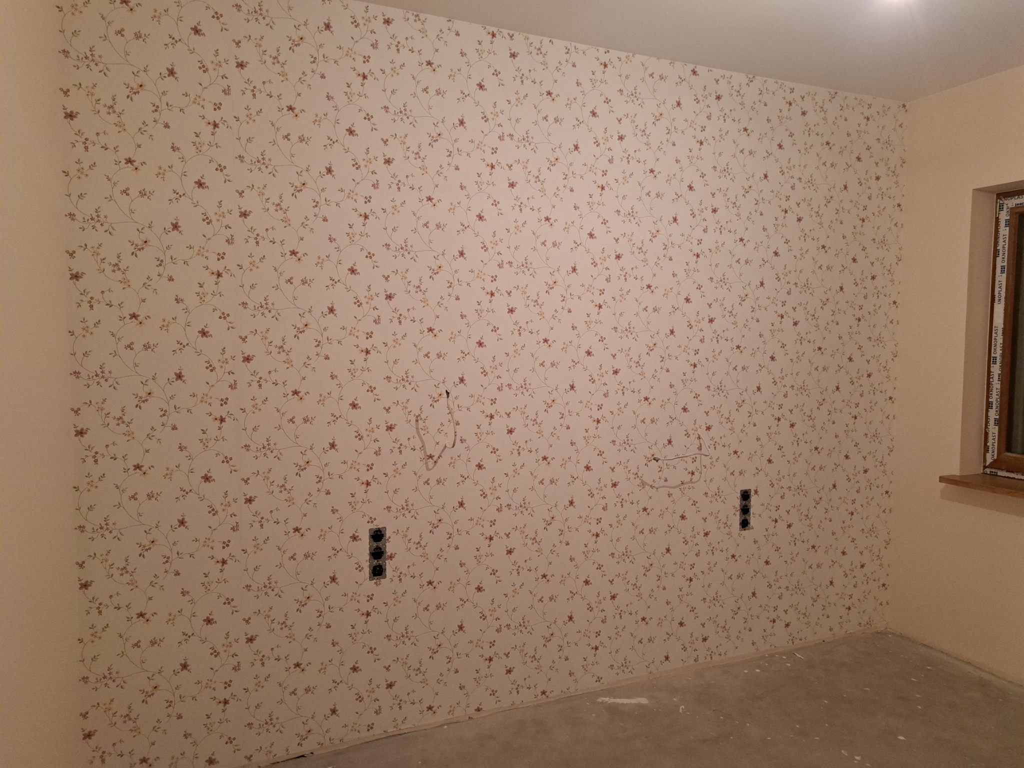 Ściana pokryta tapetą w drobne kwiatowe wzory, widoczne gniazdka elektryczne i wystające przewody, surowy beton na podłodze, okno z drewnianą ramą.