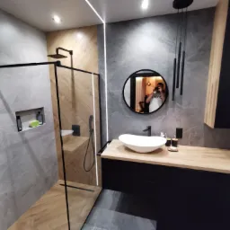 Nowoczesna łazienka z kabiną prysznicową typu walk-in, umywalką nablatową na szafce z drewnianym blatem i okrągłym lustrem, w którym odbija się osoba robiąca zdjęcie.