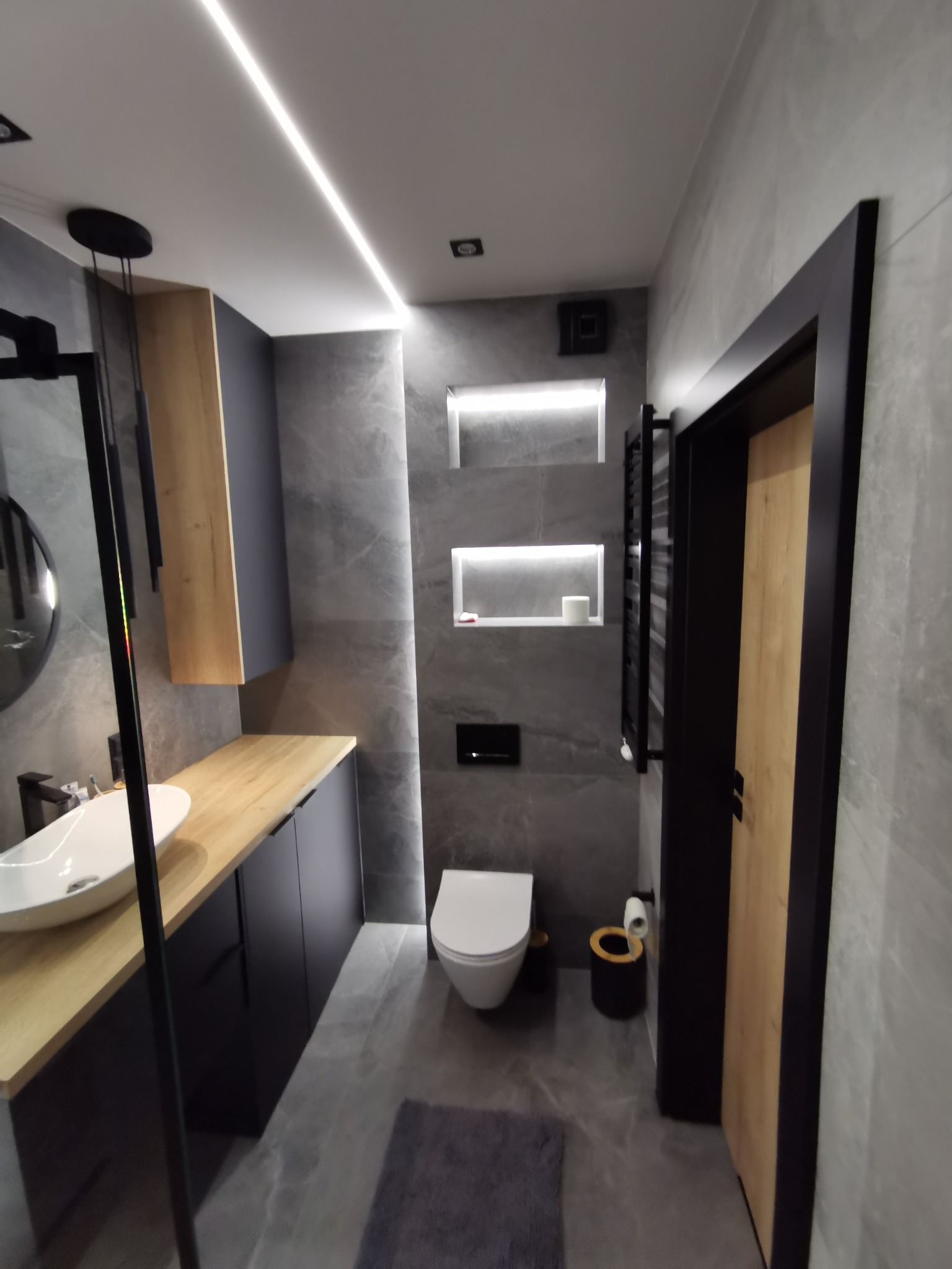 Nowoczesna łazienka w szarościach z drewnianymi akcentami: podświetlane półki, umywalka nablatowa, czarne dodatki i listwa LED na suficie.