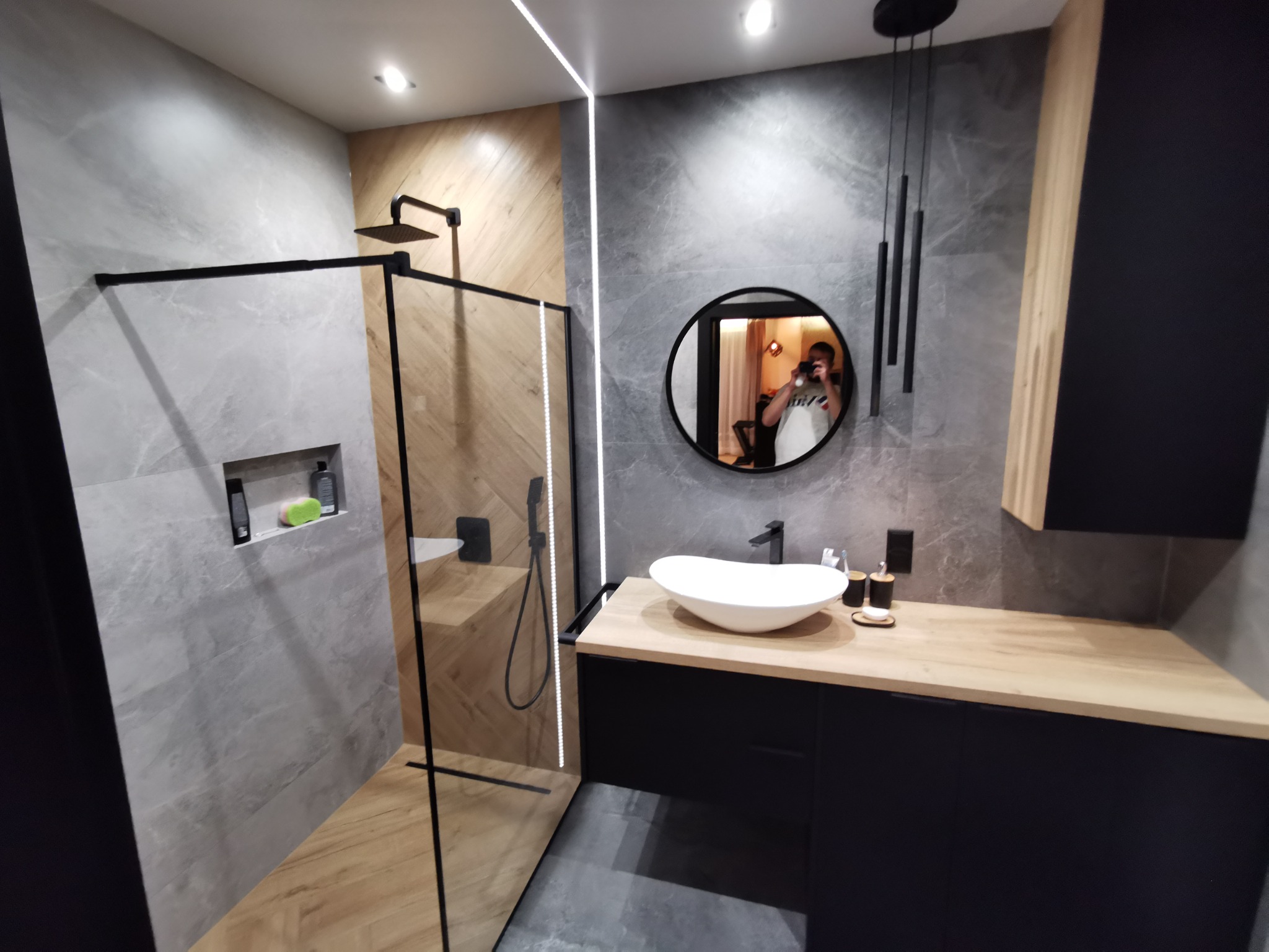 Nowoczesna łazienka z kabiną prysznicową typu walk-in, umywalką nablatową na szafce z drewnianym blatem i okrągłym lustrem, w którym odbija się osoba robiąca zdjęcie.
