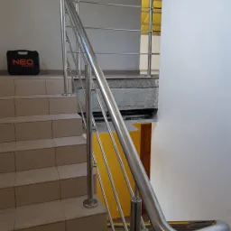 Schody z beżowych płytek z zamontowaną, nowoczesną balustradą ze stali nierdzewnej z poziomymi wypełnieniami; na podeście stoi czarna skrzynka narzędziowa marki NEO Tools.