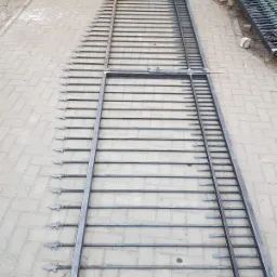 Surowa, metalowa brama wjazdowa z ostrymi zdobieniami na szczytach, leżąca na brukowanej nawierzchni w trakcie spawania, z widocznymi śladami po spawaniu.