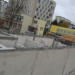 Budowa fundamentów z bloczków betonowych, widoczne zbrojenia i stalowe łączniki, w tle budynki miejskie i reklama szkoły jazdy.