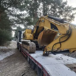 Żółta koparka gąsienicowa na niskopodwoziowej platformie transportowej, z widocznymi śladami błota i śniegu, przewożona leśną drogą.