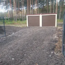 Dwu-stanowiskowy garaż z brązowymi bramami i beżowymi obramowaniami, na nierównym, przygotowanym terenie, w tle wysoki las sosnowy.