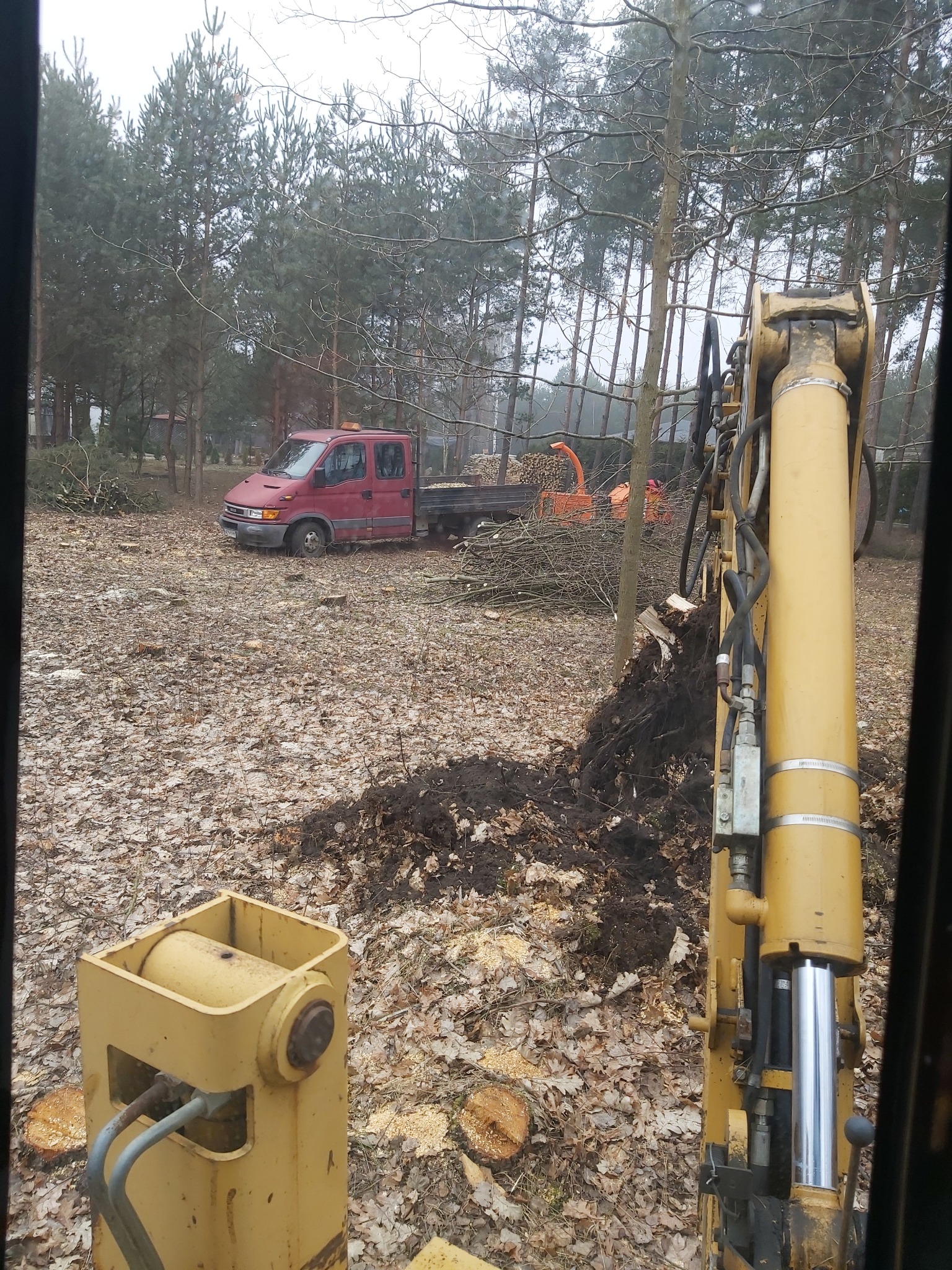 Widok z kabiny maszyny budowlanej na czerwony samochód dostawczy z przyczepą wypełnioną gałęziami i rozdrabniarką do gałęzi na tle lasu.