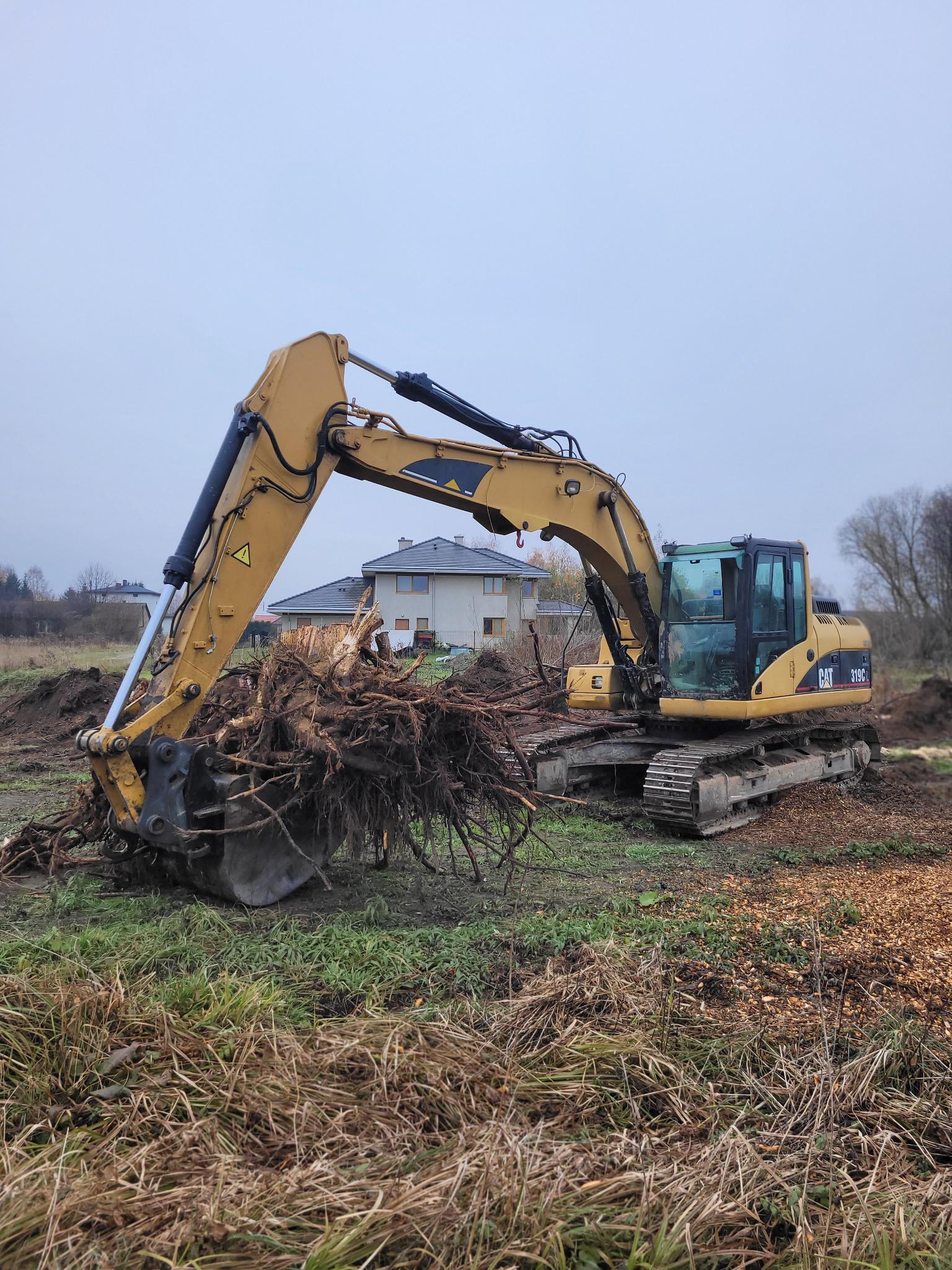 Żółta koparka CAT 319C z pełną łyżką korzeni drzew na terenie budowy z widocznym domem w tle, pochmurne niebo.