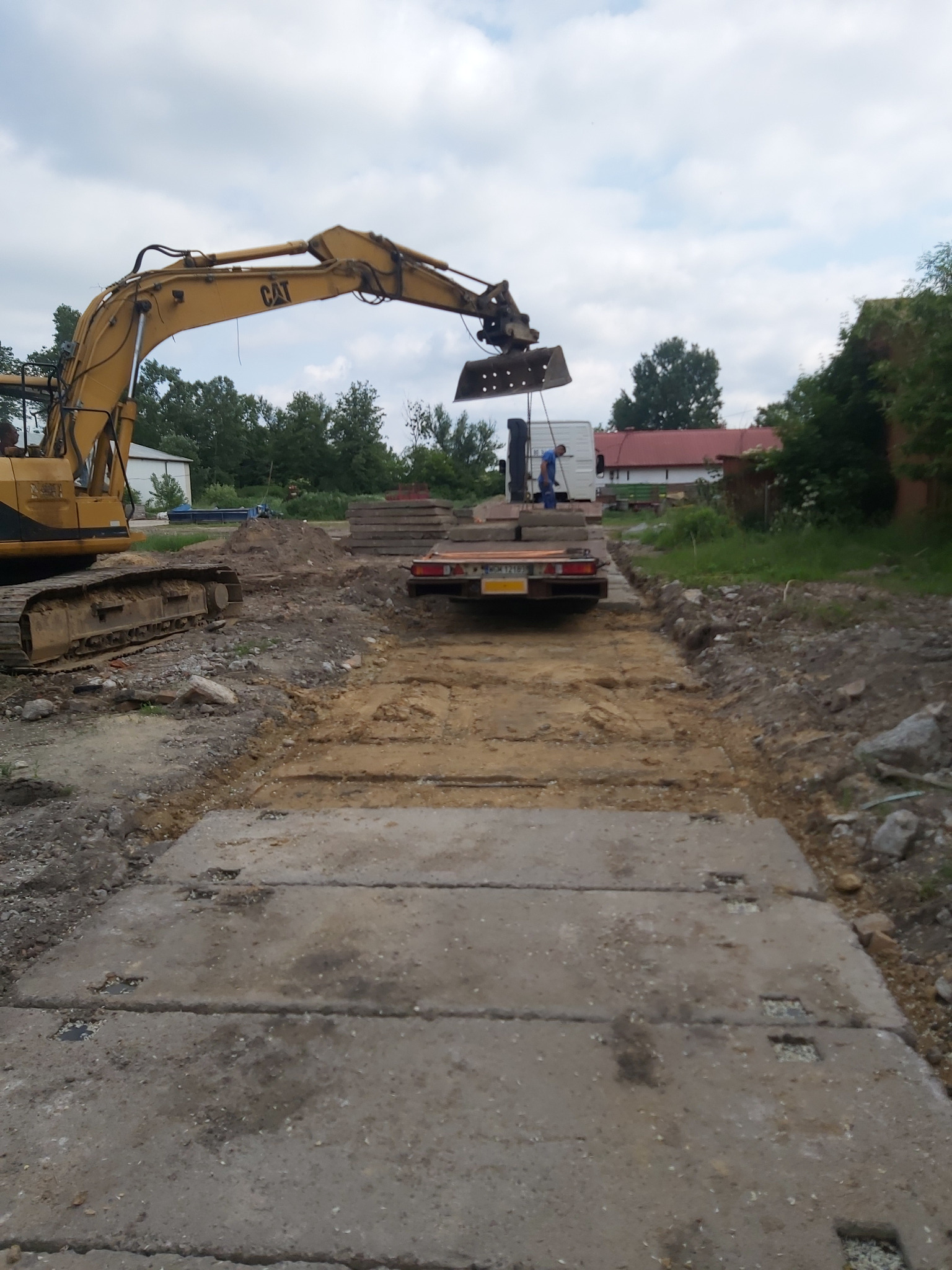 Żółta koparka CAT umieszcza betonowe płyty na przygotowanym podłożu, widoczna naczepa z kolejnymi płytami, w tle budynek z czerwonym dachem.