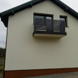 Elewacja budynku z beżową ścianą, brązowym cokołem i czarnymi ramami okiennymi oraz balkonem z poziomymi barierkami, widok z boku w pochmurny dzień.