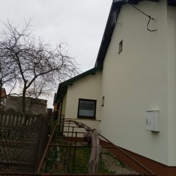 Branz-Bud - Świeżo otynkowana ściana budynku z widoczną skrzynką elektryczną i rurą spustową, w tle drzewo bez liści i gruzowisko.