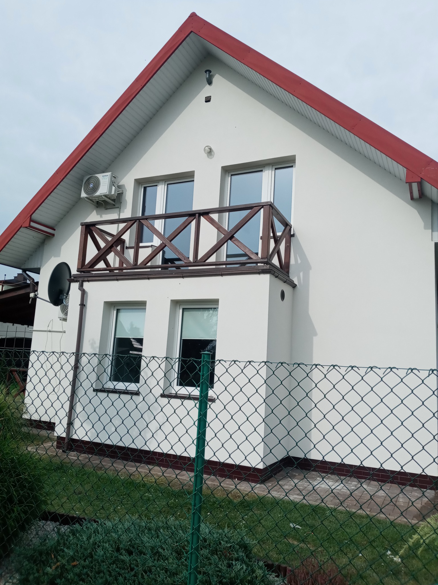 Dom jednorodzinny z białą elewacją, czerwonym dachem i drewnianą balustradą balkonu, widoczny zza zielonego ogrodzenia z siatki.