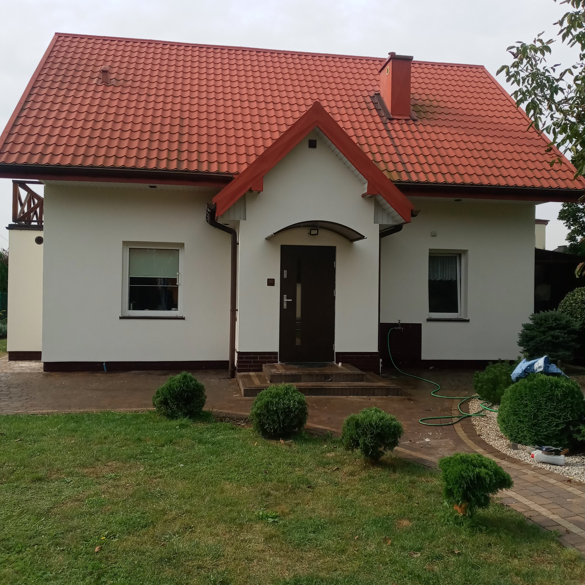 Ocieplony dom jednorodzinny z czerwoną dachówką, widoczne świeżo otynkowane białe elewacje, zielony trawnik i ozdobne krzewy, wężyk ogrodowy leżący obok.