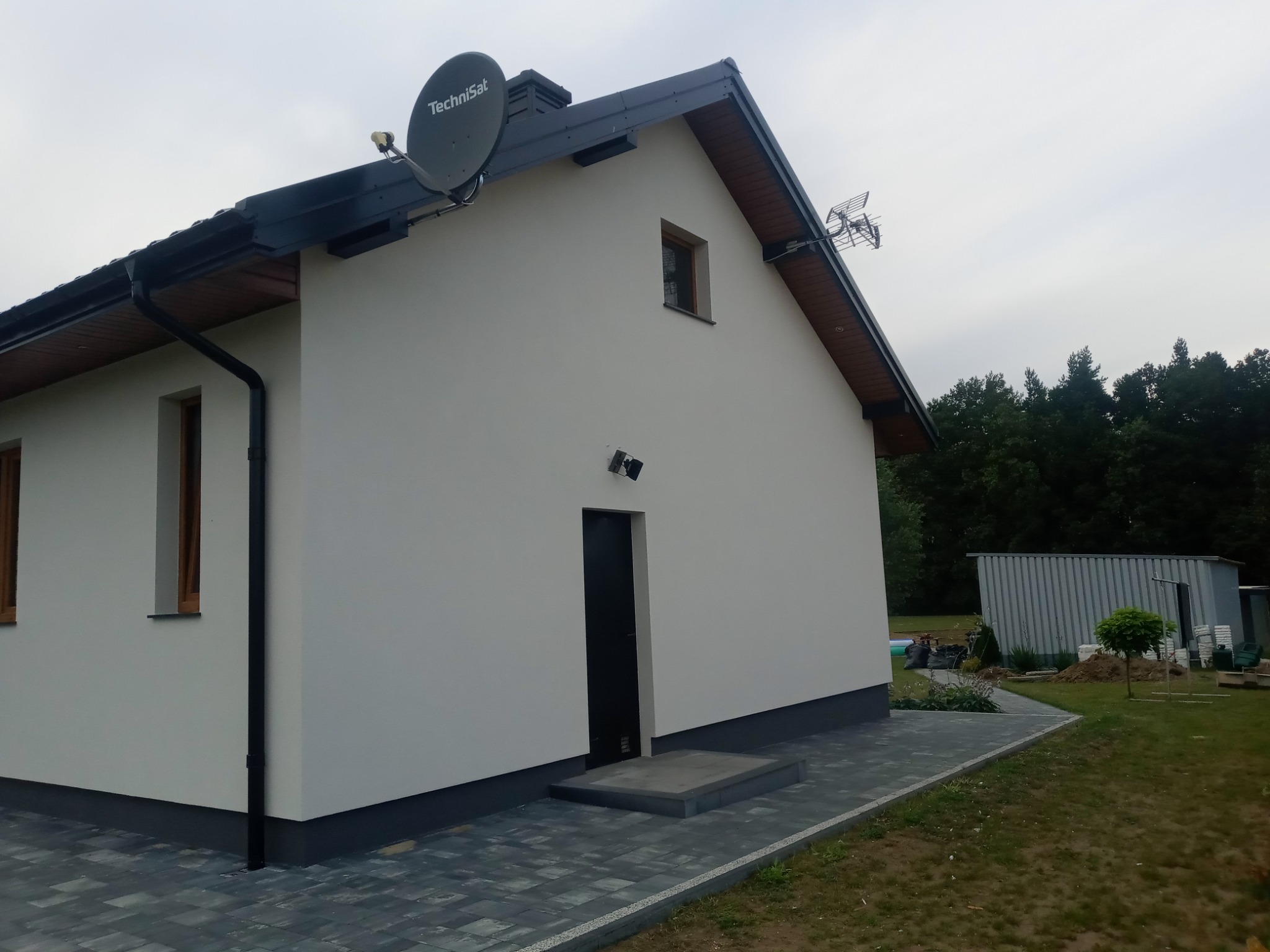 Wykończony dom jednorodzinny z jasną elewacją, ciemną podbitką i dachem, widoczna antena satelitarna TechniSat i antena naziemna, szara kostka brukowa wokół budynku.