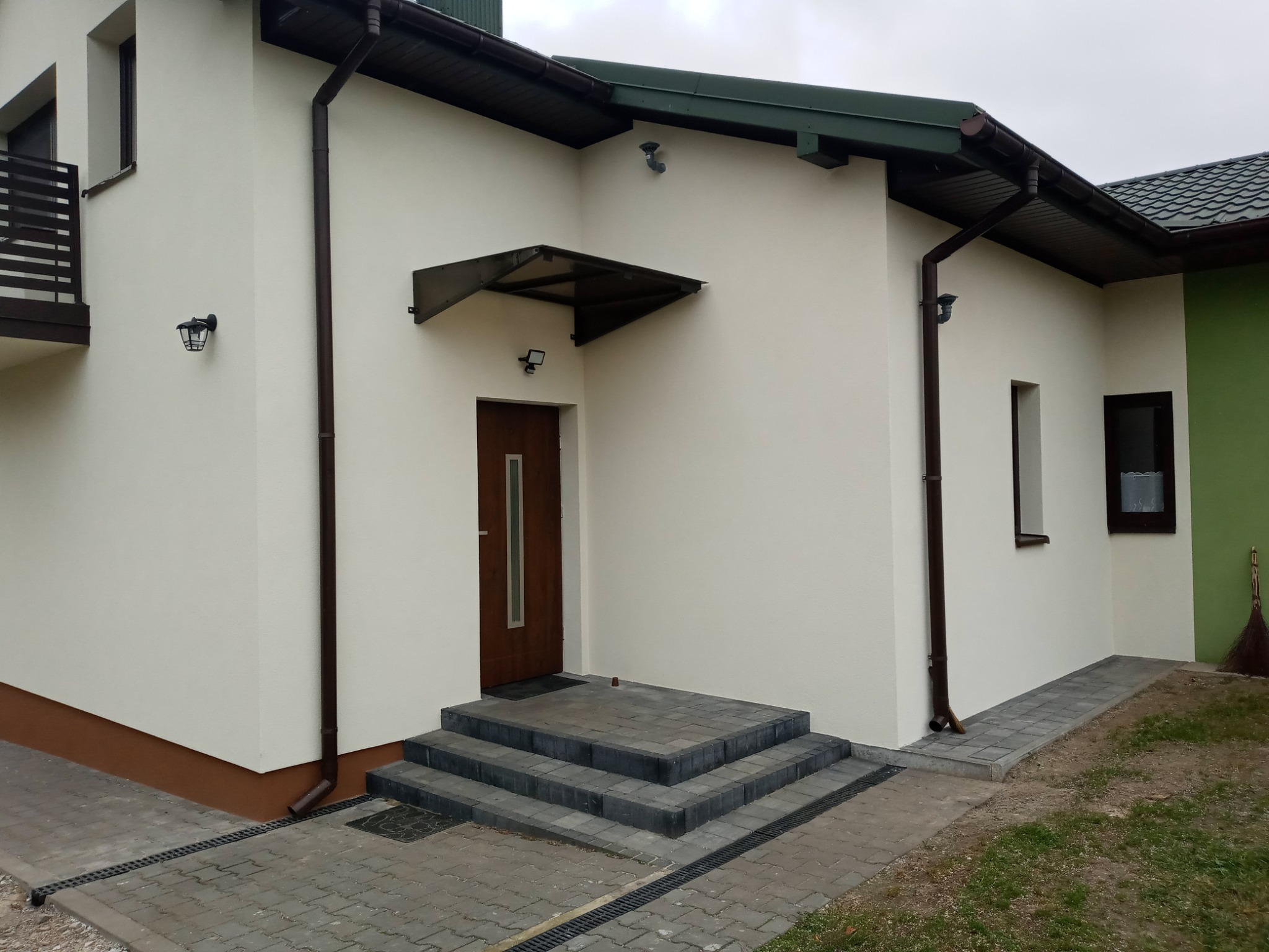 Wykończona elewacja domu jednorodzinnego z brązowymi rynnami, ciemnobrązowym dachem i schodami wejściowymi z szarej kostki brukowej.