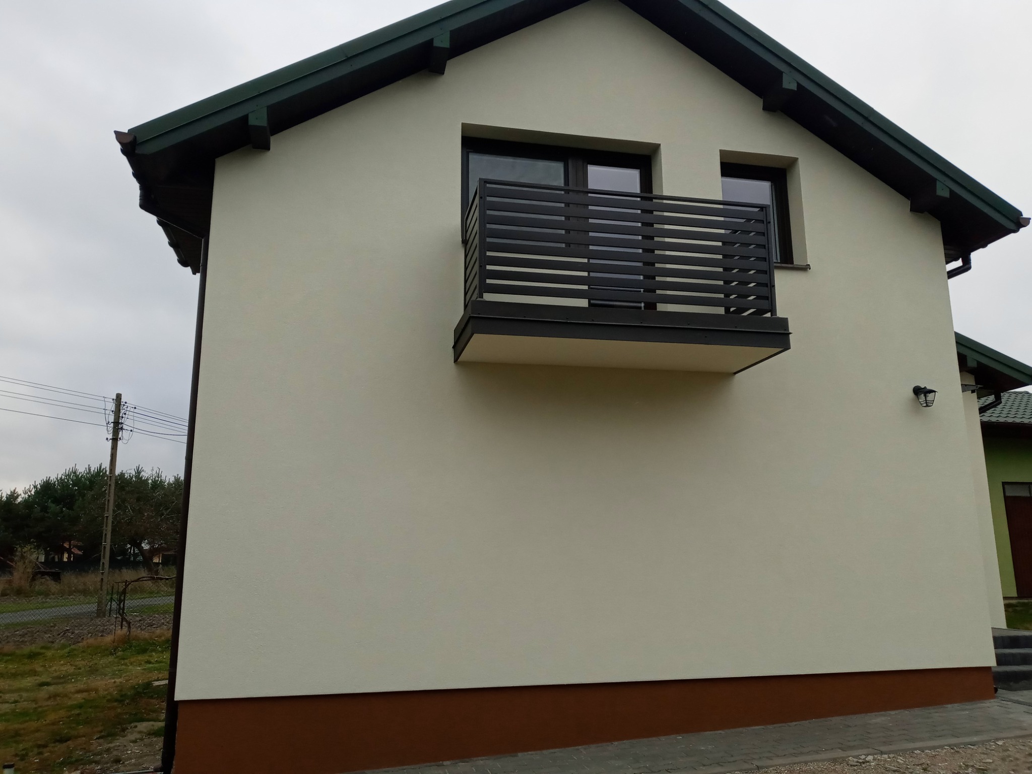 Elewacja budynku z beżową ścianą, brązowym cokołem i czarnymi ramami okiennymi oraz balkonem z poziomymi barierkami, widok z boku w pochmurny dzień.