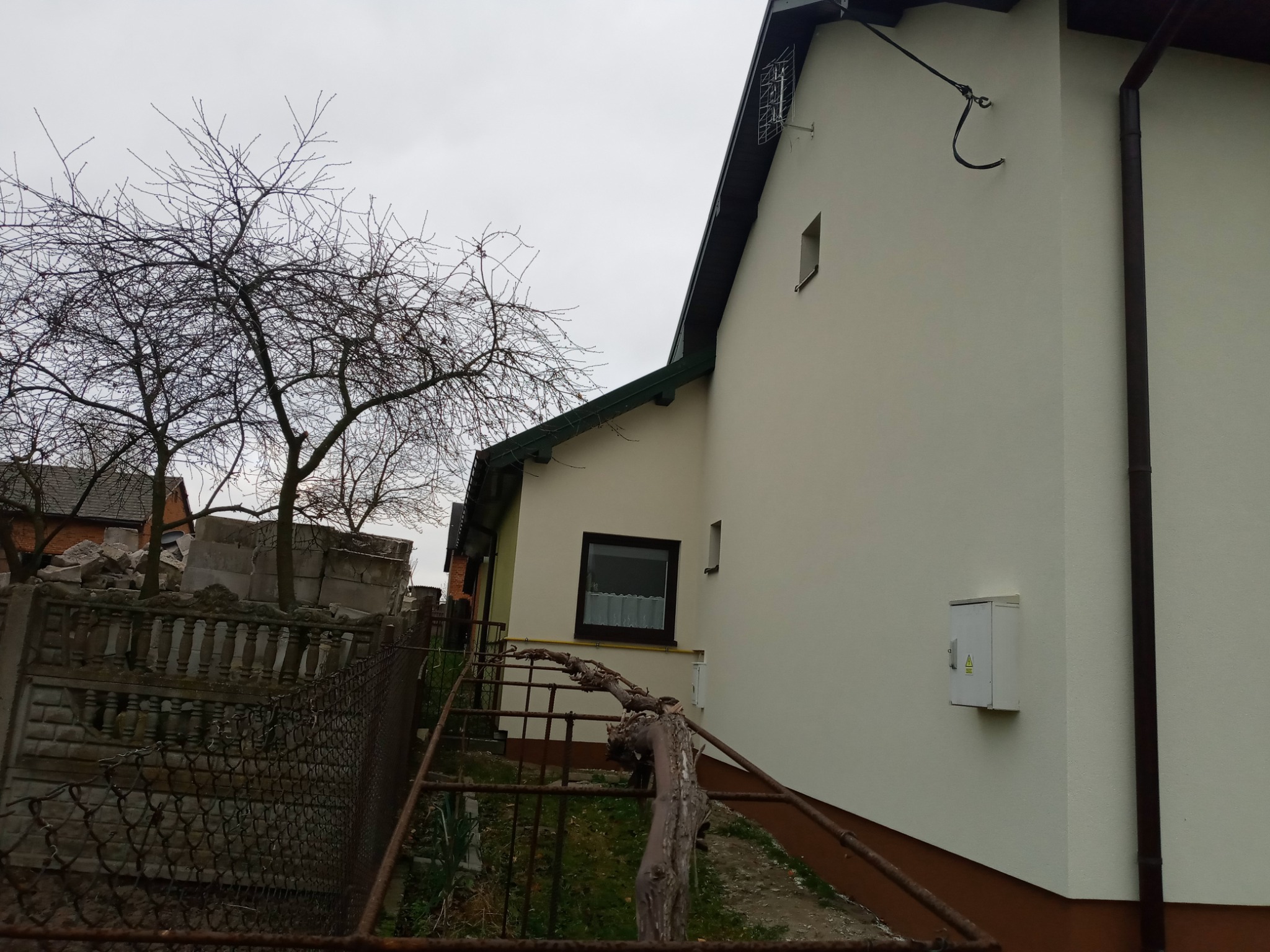 Świeżo otynkowana ściana budynku z widoczną skrzynką elektryczną i rurą spustową, w tle drzewo bez liści i gruzowisko.