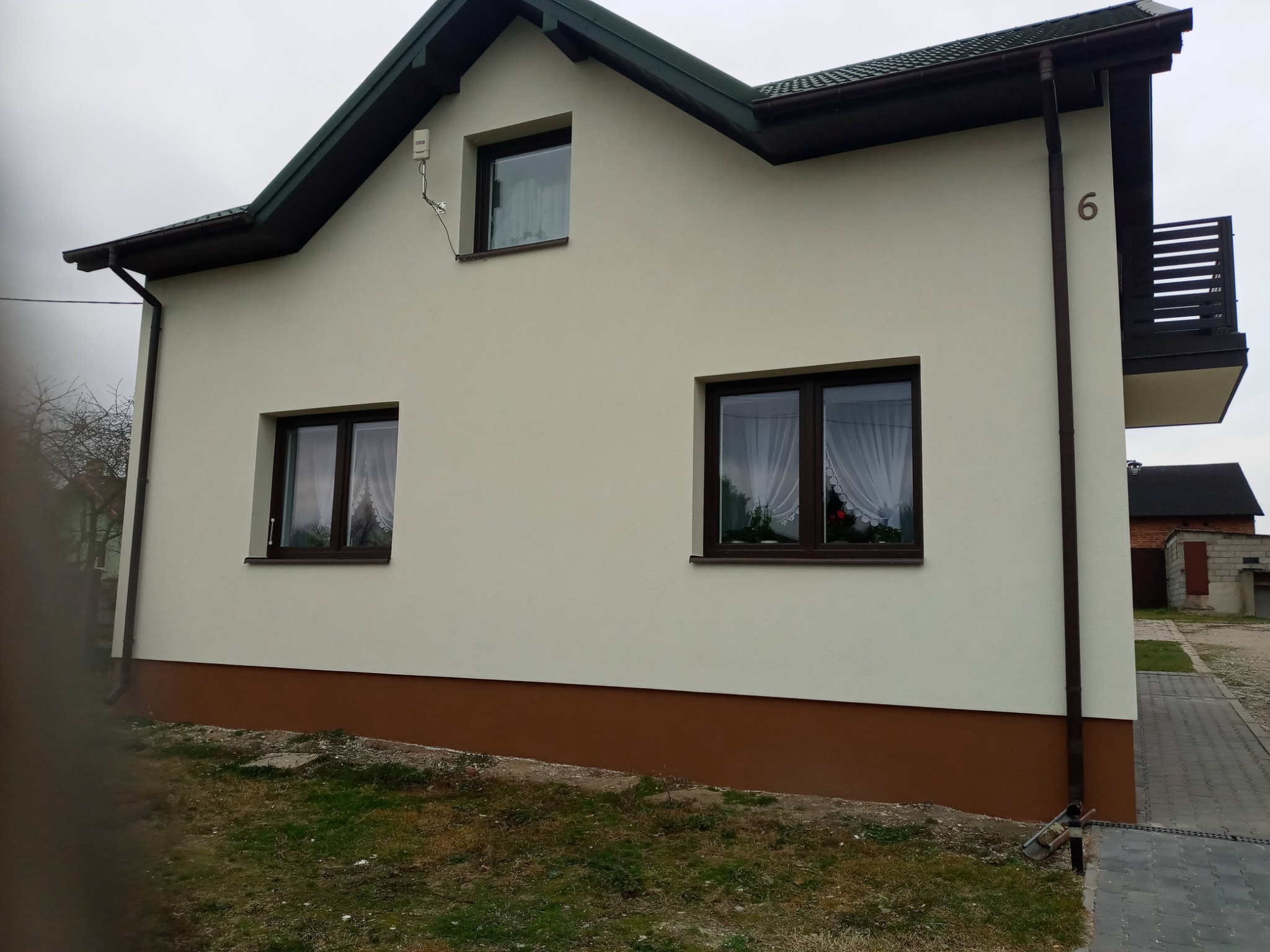 Ocieplona elewacja domu jednorodzinnego z brązowym cokołem, trzema oknami z firankami i numerem '6' na ścianie.