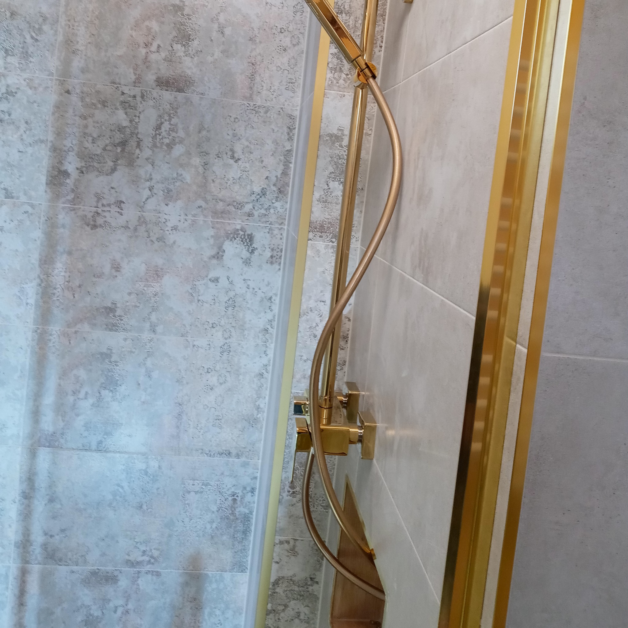 Nowoczesna kabina prysznicowa z deszczownicą i złotymi elementami wykończeniowymi, szklane drzwi, płytki imitujące beton na ścianach.