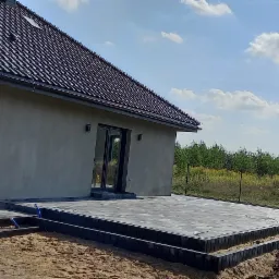 Nowoczesny taras z ciemnoszarej kostki brukowej, obramowany obrzeżami, przylegający do szarego domu z ciemną dachówką, widok na ogród w tle, częściowo przygotowany teren pod dalsze prace.