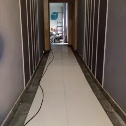 Korytarz w trakcie prac wykończeniowych: świeżo ułożone płytki podłogowe w odcieniach bieli, ściany pomalowane na szaro z pionowymi białymi pasami, widoczny kabel i wiadro z zaprawą, w głębi...