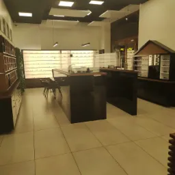 Wnętrze salonu optycznego z ekspozycją okularów na regałach i wyspie sprzedażowej z krzesłami, oświetlone panelami LED.