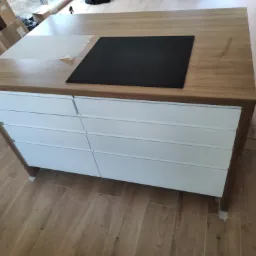 Blat drewniany z wbudowaną płytą indukcyjną i białymi frontami szuflad, widoczne kartony w tle, całość na podłodze imitującej drewno.