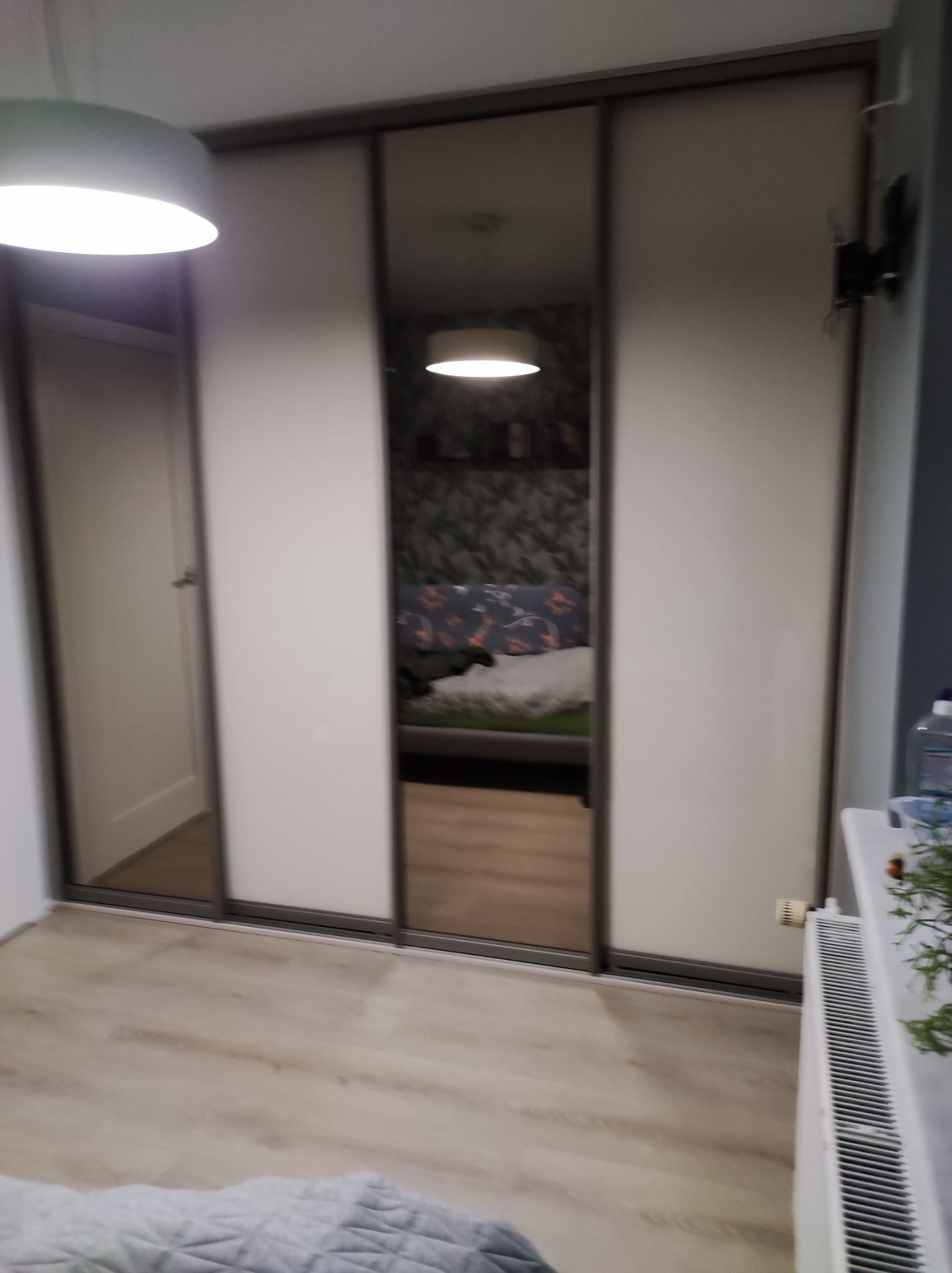 Trzydrzwiowa szafa przesuwna z lustrzanym środkowym panelem, odbijającym wystrój sypialni z wzorzystą tapetą i łóżkiem w tle.