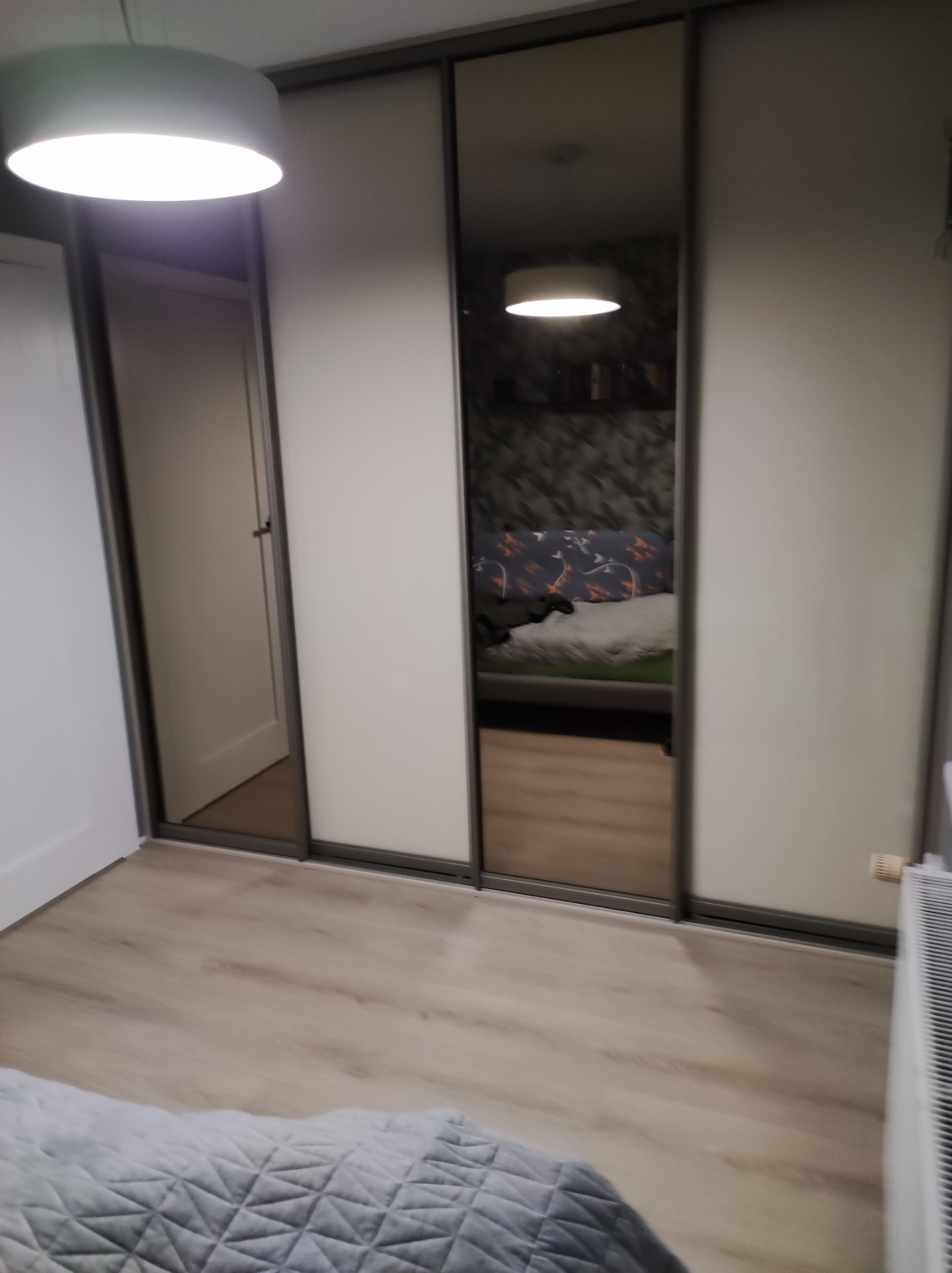 Szafa wnękowa z przesuwnymi drzwiami, częściowo lustrzanymi, odbijająca fragment sypialni z wzorzystą tapetą i łóżkiem.