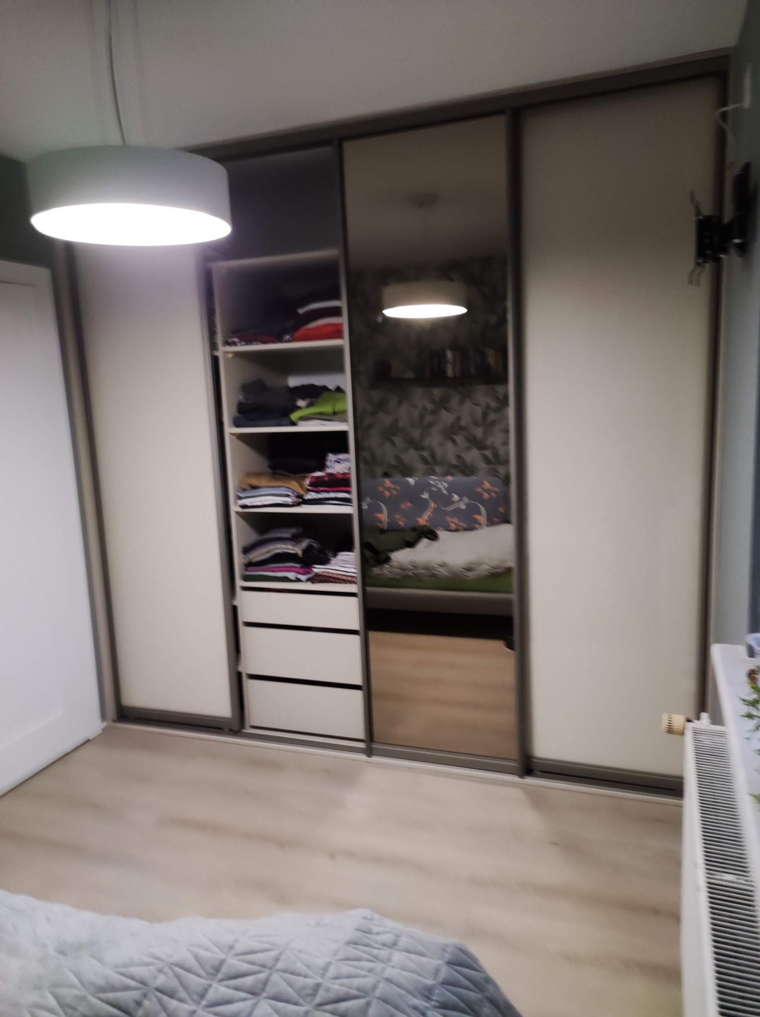 Trzydrzwiowa szafa przesuwna z lustrem pośrodku, z widoczną zawartością półek i szuflad po lewej stronie, odbicie pokoju w lustrze.