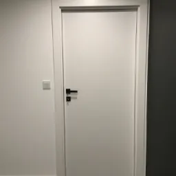 Białe drzwi wewnętrzne z czarną klamką, osadzone w białej ościeżnicy, na tle białej ściany z włącznikiem światła i ciemnej ściany po prawej stronie.