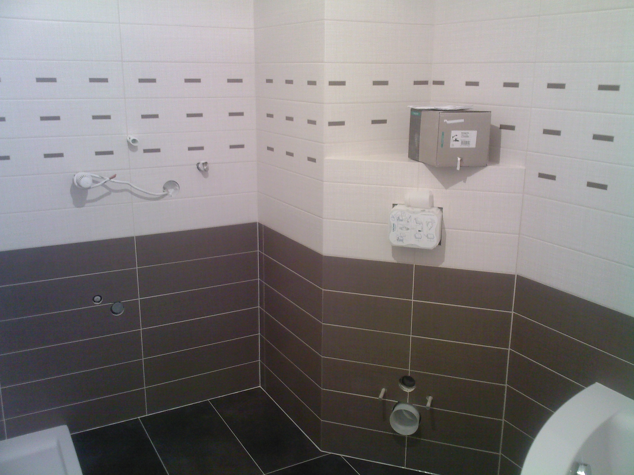 Wnętrze łazienki w trakcie remontu z widocznymi białymi i brązowymi płytkami, instalacją wodną i pudełkiem na ścianie.