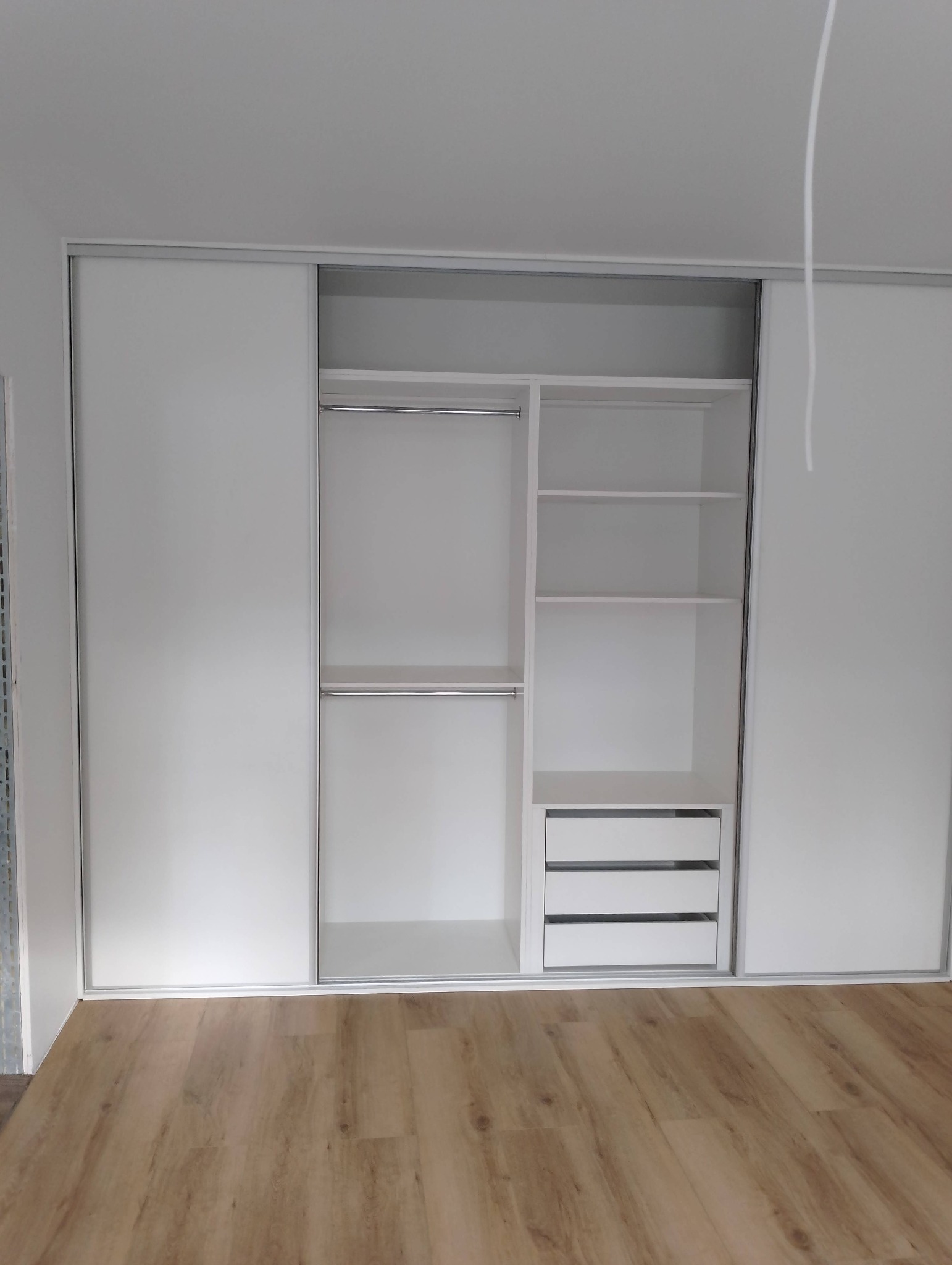 Biała szafa wnękowa z przesuwnymi drzwiami, wewnątrz drążki na ubrania, półki i trzy szuflady, na podłodze panele imitujące drewno.