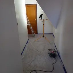 Pokój na poddaszu w trakcie malowania: świeżo pomalowane ściany na biało, zabezpieczona folią podłoga, drabina oparta o drewniane drzwi, reflektor na trójnogu oświetla pomieszczenie.