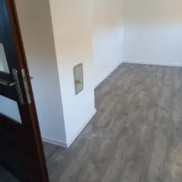 Pusty pokój po remoncie z szarą podłogą imitującą drewno, białe ściany z listwami przypodłogowymi i metalową skrytką z kluczem, widok zza otwartych, ciemnych drzwi.