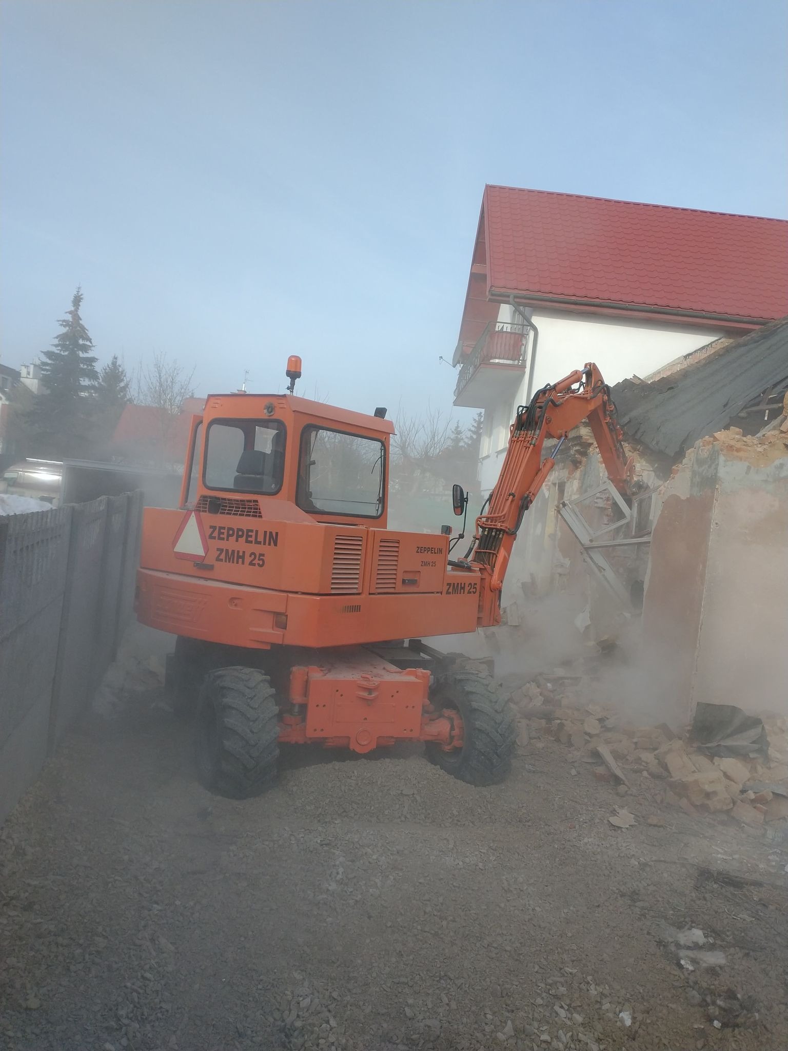 Pomarańczowa koparka Zeppelin ZMH 25 podczas prac rozbiórkowych, unoszący się pył, w tle budynek z czerwonym dachem.