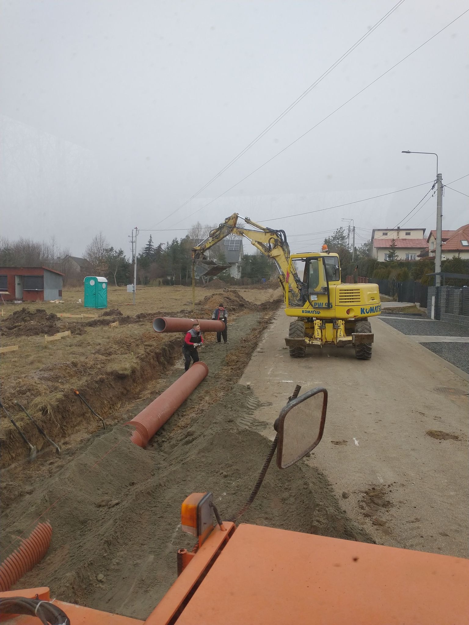 Żółta koparka Komatsu na placu budowy, robotnicy układają rury kanalizacyjne w wykopie, w tle widoczny przenośny toi-toi i częściowo ogrodzenie.