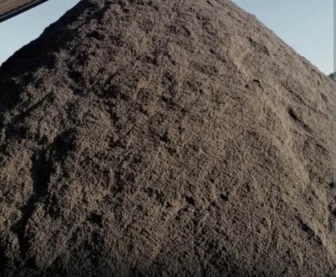 Góra ciemnego, drobnego kruszywa, prawdopodobnie ziemi lub piasku, usypana w stożek, z widocznym fragmentem maszyny budowlanej w górnym rogu.