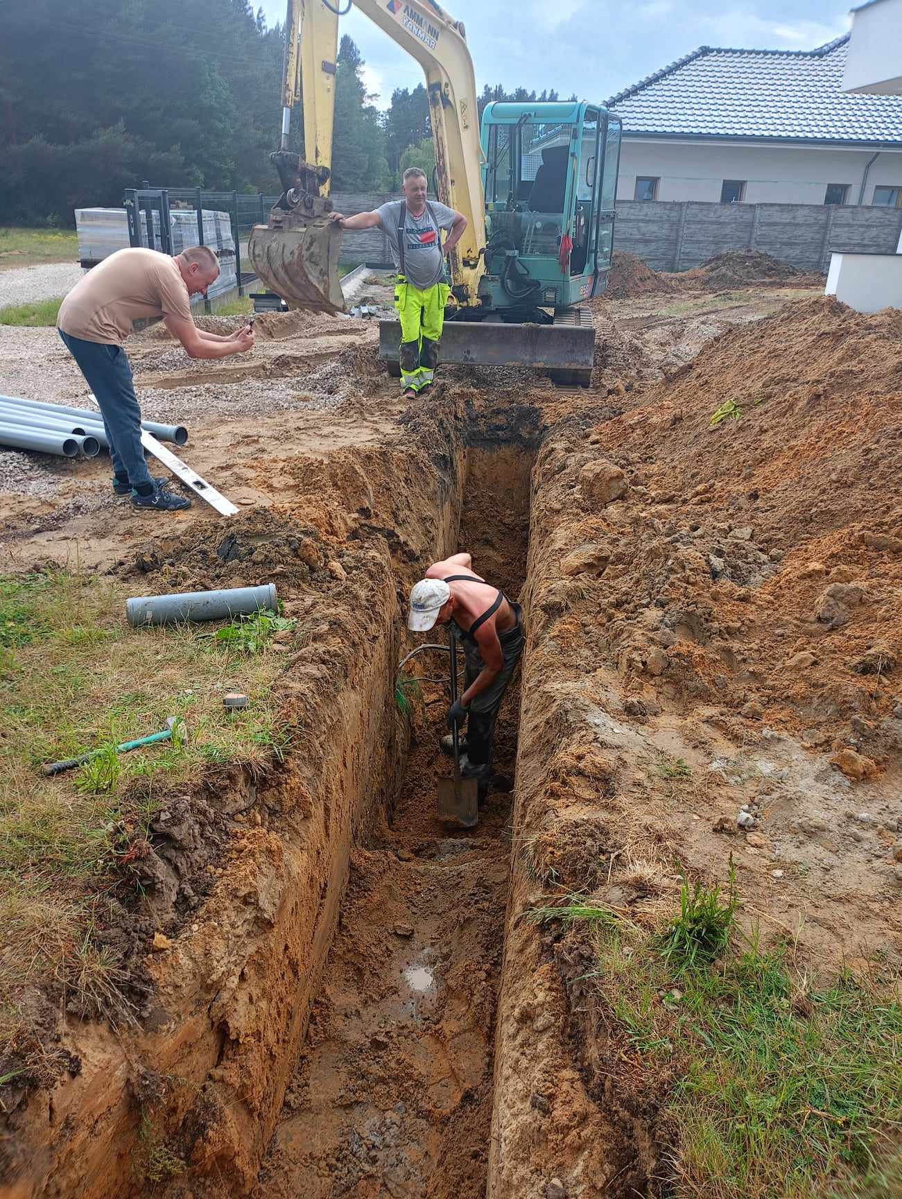 Głęboki wykop liniowy w ziemi, pracownik z łopatą na dnie, rury kanalizacyjne obok, operator koparki nadzoruje, drugi pracownik robi zdjęcie telefonem, widoczny dom w tle.