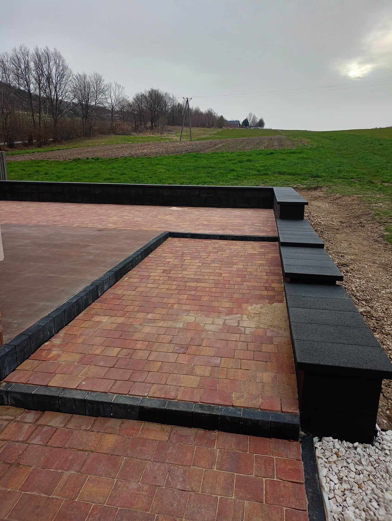Ułożona czerwono-pomarańczowa kostka brukowa z czarnym obramowaniem na tle zielonego pola i pochmurnego nieba. Widoczne schodki z kostki i fragment białego żwiru.