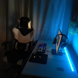 Ergonomiczne czarno-białe krzesło gamingowe GTR ustawione przy biurku z podświetleniem LED, klawiaturą, myszką i słuchawkami na tle zasłony.