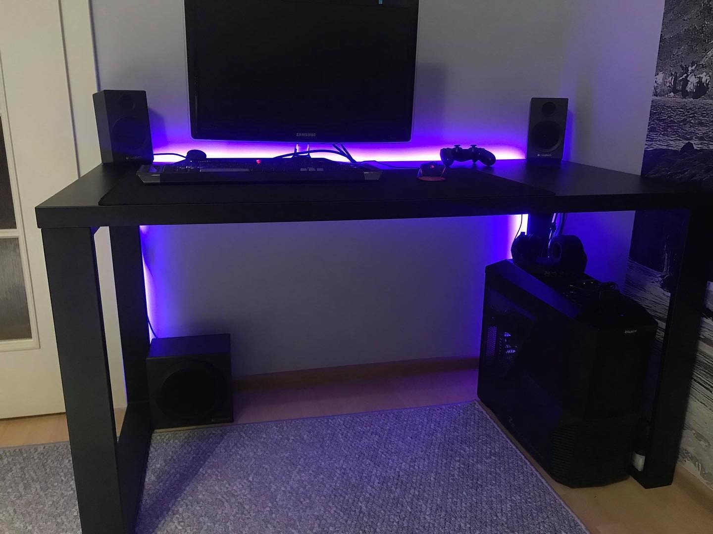 Czarne biurko gamingowe z podświetleniem LED, monitor, głośniki, klawiatura, mysz i komputer stacjonarny widoczne na blacie i pod nim.