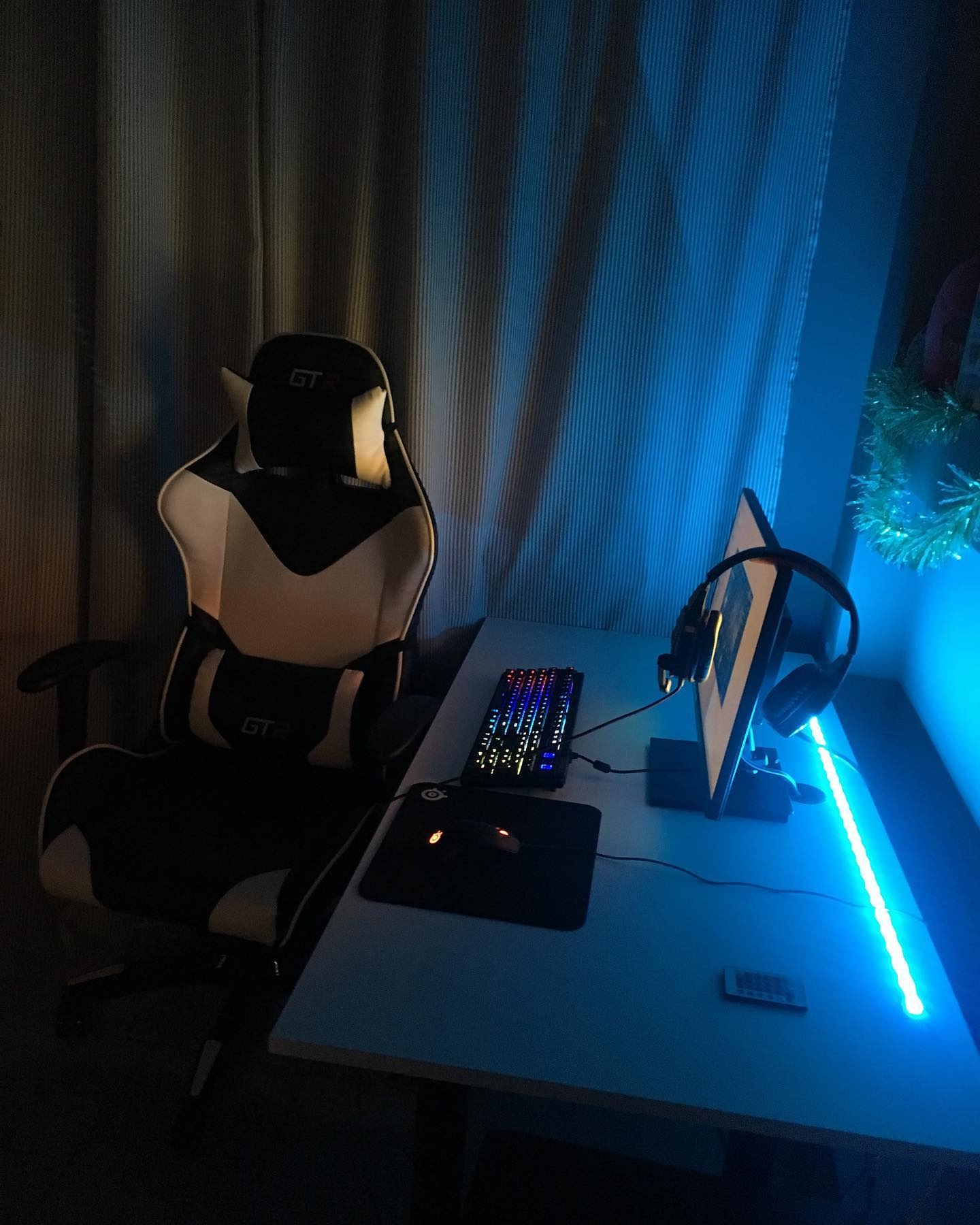 Ergonomiczne czarno-białe krzesło gamingowe GTR ustawione przy biurku z podświetleniem LED, klawiaturą, myszką i słuchawkami na tle zasłony.
