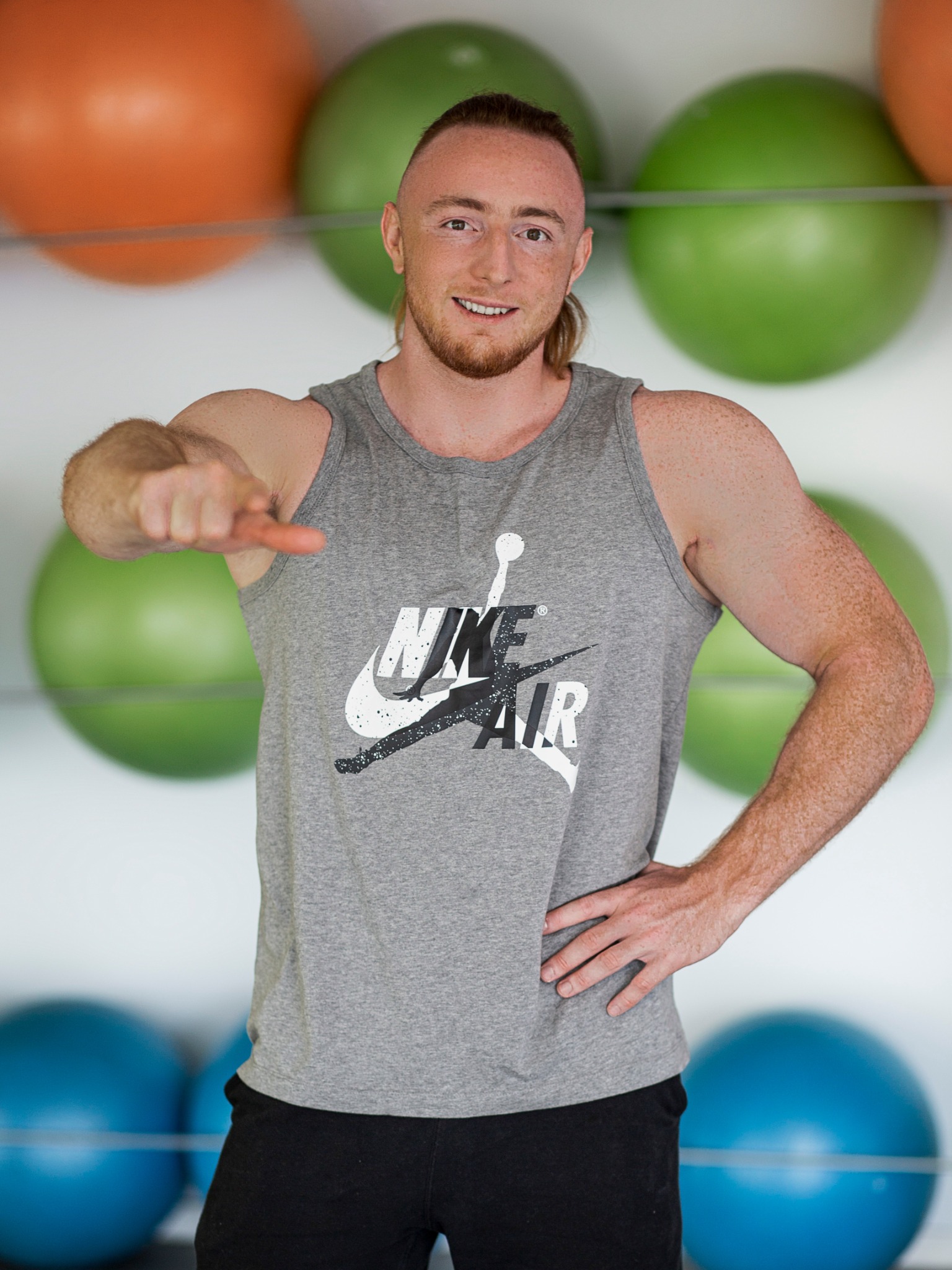 Uśmiechnięty mężczyzna w sportowej koszulce bez rękawów, stojący na tle kolorowych piłek do ćwiczeń, wyciąga palec wskazujący w kierunku widza.