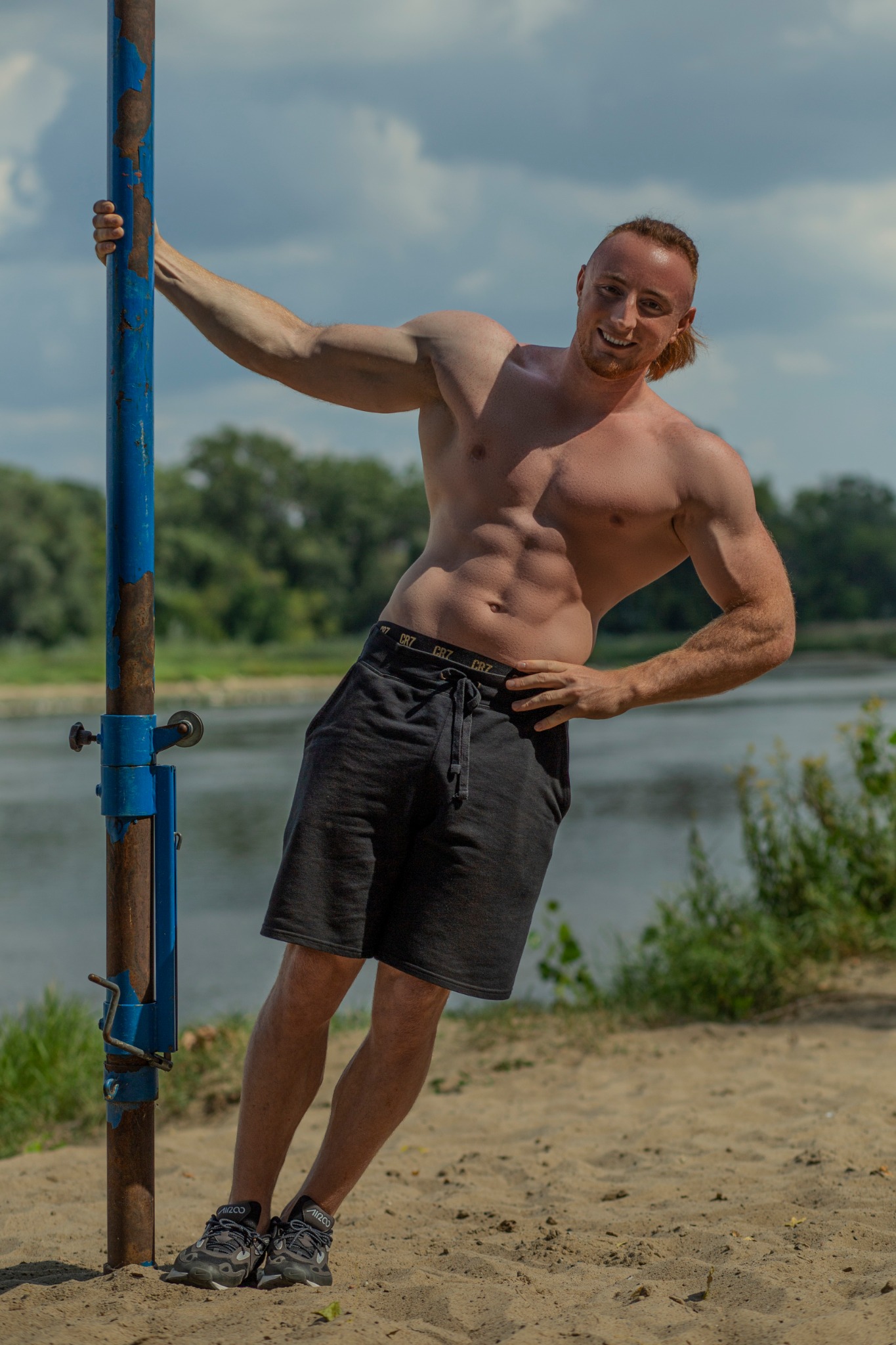 Uśmiechnięty, wysportowany mężczyzna w czarnych szortach i sportowych butach, oparty o niebieski, częściowo zardzewiały słup na piaszczystej plaży z widokiem na rzekę i drzewa w tle.