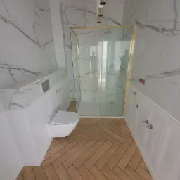 Nowoczesna łazienka z marmurowymi płytkami na ścianach, drewnianą podłogą ułożoną w jodełkę, podwieszanym WC i kabiną prysznicową ze złotymi ramami.