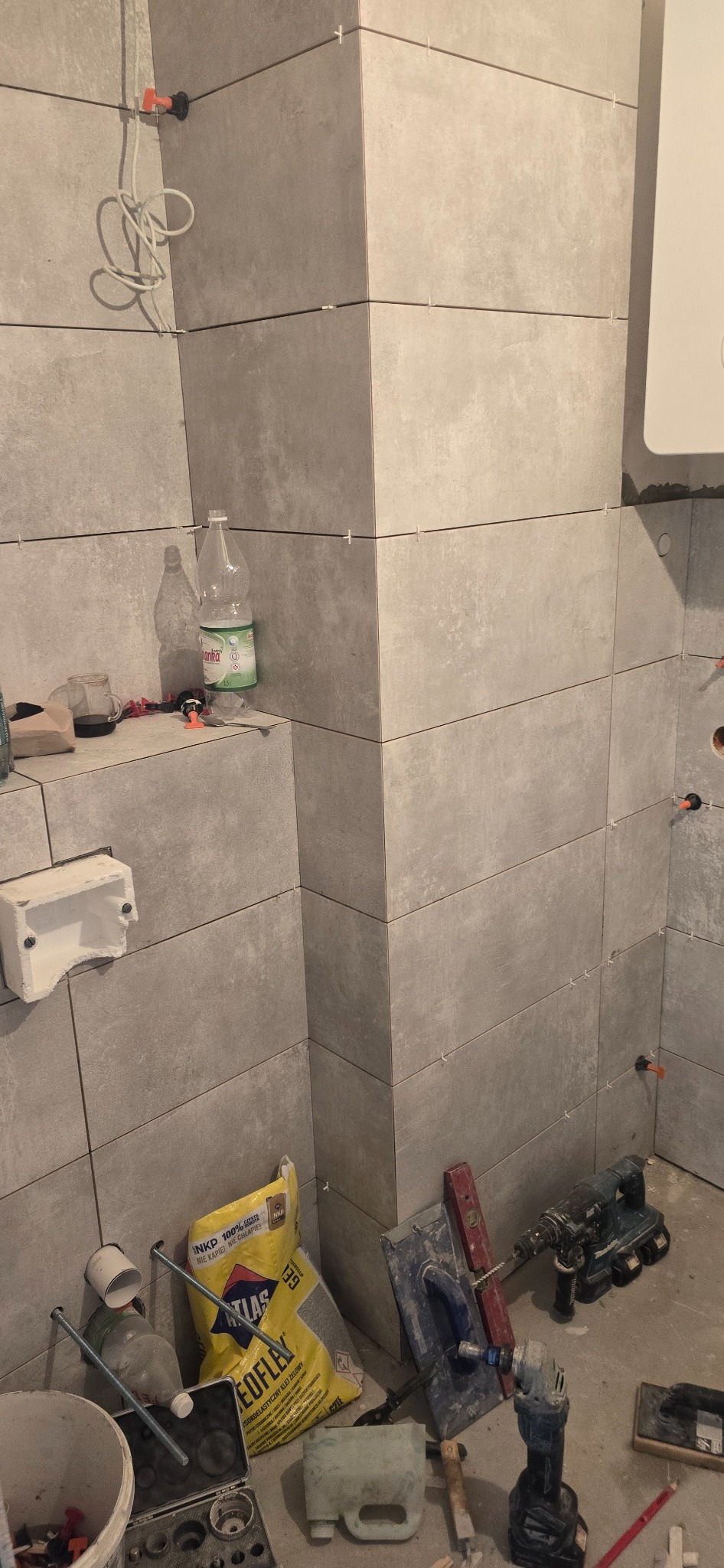 Świeżo ułożone, szare, matowe płytki na ścianie w łazience, z widocznymi krzyżykami dystansowymi, workiem zaprawy klejowej Atlas Geoflex, narzędziami glazurniczymi i butelką wody mineralnej w tle.