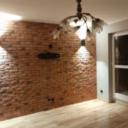 Salon z ceglaną ścianą, oświetleniem punktowym i drewnianą podłogą, widok na okno z grzejnikiem.