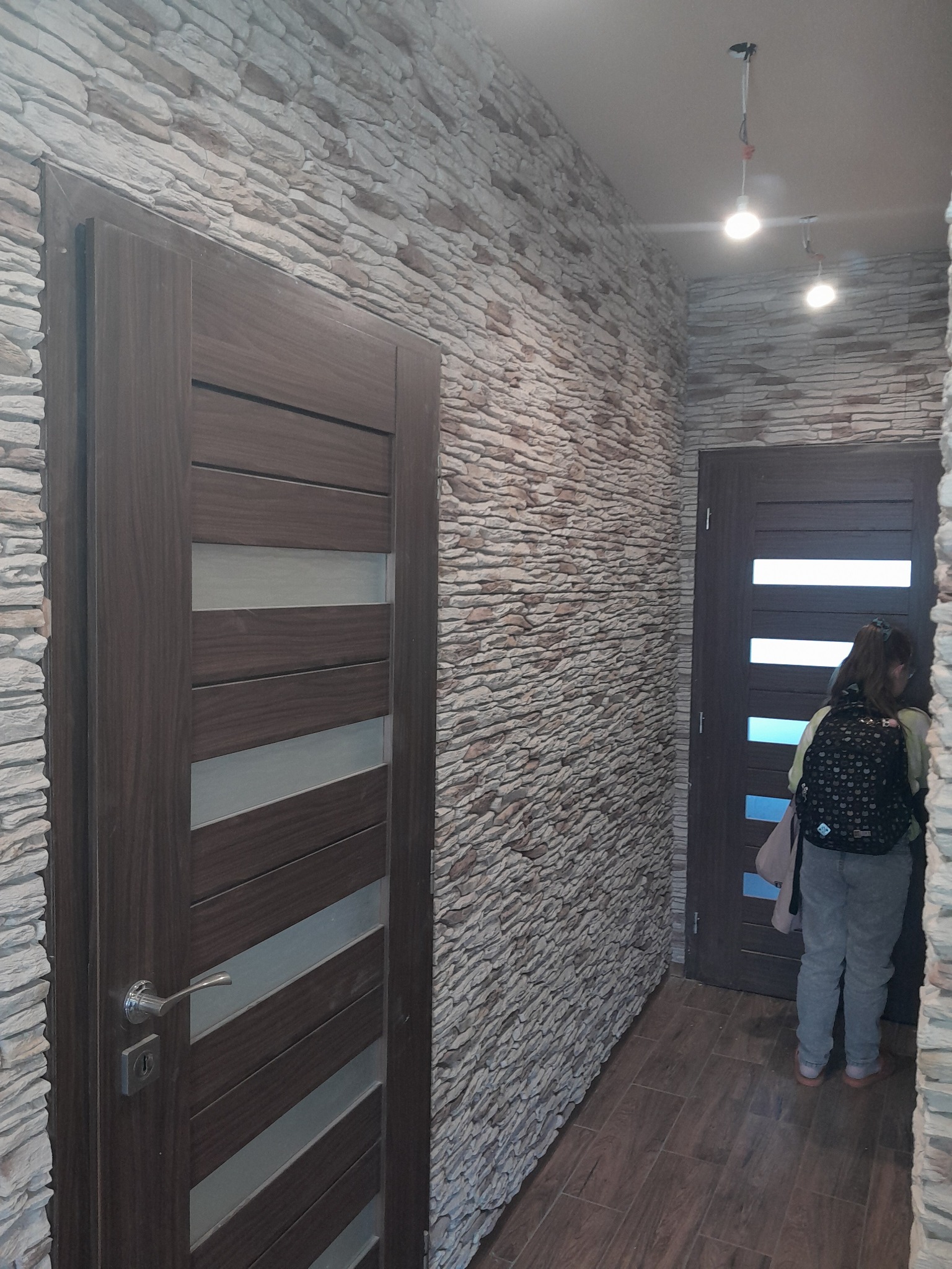 Wnętrze korytarza z kamienną okładziną ściany, drewnianą podłogą i dwoma ciemnymi drzwiami z poziomymi wstawkami, jedno z nich otwarte. Widoczna osoba z plecakiem wchodząca do pomieszczenia.
